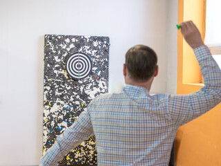 A man viewed from behind throws a green dart toward a black and white circular target. The target is mounted on a speckled foam backstop against a white wall. selective focus
