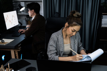 Businesswoman with stressful face looking document paperwork file while coworker trading stock market at back at over late night time at modern office, working on startup company project. Infobahn.
