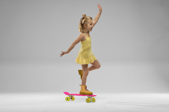 small blonde girl balances on a skateboard while wearing a bright dress and yellow boots. She has a joyful expression and raises one hand in excitement. - Powered by Adobe