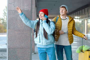Young couple with baggage talking by mobile phone near airport on autumn day