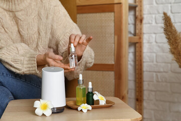 Woman with air humidifier, essential oils and plumeria flowers on table in room, closeup