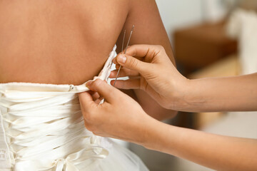 Woman with needles fitting wedding dress of beautiful young bride at home, back view