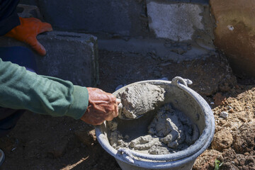 Obraz premium Focused manual labor on construction site. person wearing glove mixes gray cement mortar in bucket for foundation repair, showing hard work and skilled effort