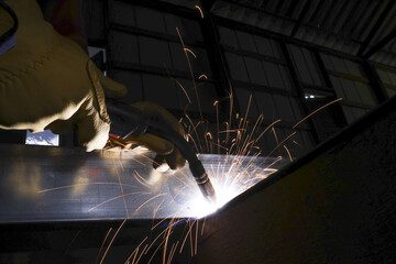 Focused welder at work in dark industrial factory. Wearing protective glove while welding metal, creating shower of bright, hot sparks during dangerous job