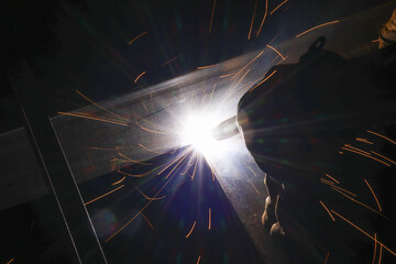 Focused industrial welder at work, welding metal with bright light and hot sparks. This construction job shows intense fabrication, skilled manual worker in dark workshop