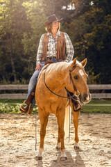 Obraz premium Best ager woman sitting on her palomino Quarter Horse gelding in western gear, relaxed and smiling in warm sunlight outdoors.