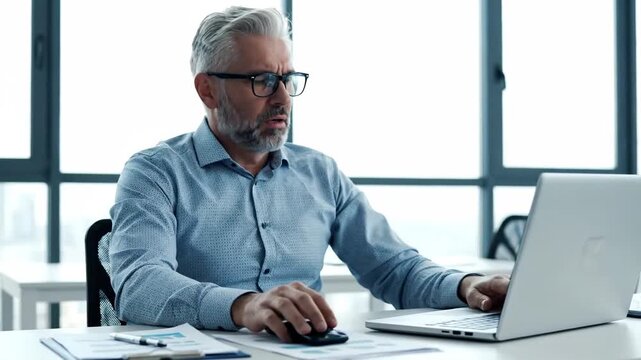 Middle aged gray haired man looking worried while working at a white desk with a computer and office documents in a light filled modern office space.