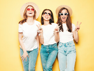 Obraz premium Three young beautiful smiling hipster female in trendy same summer white t-shirt and jeans clothes. Sexy carefree women posing near wall in studio. Cheerful and positive models having fun