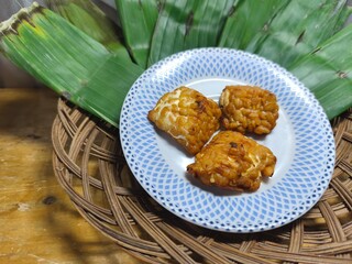 The side dish consists of three pieces of delicious fried tempeh, served on a plate decorated with banana leaves and woven rattan. It's perfect for culinary, product, hospitality, or advertising idea.