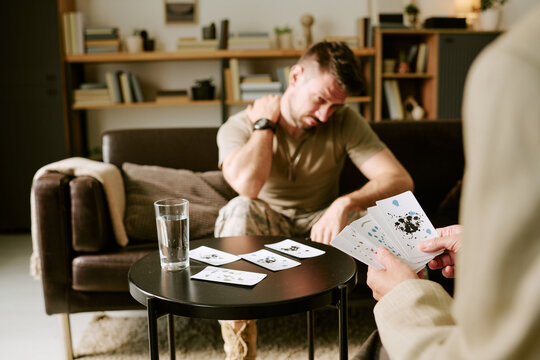 Caucasian young adult man sitting on sofa holding neck appearing distressed while another holding Rorschach inkblot cards conducting psychological assessment in therapy session