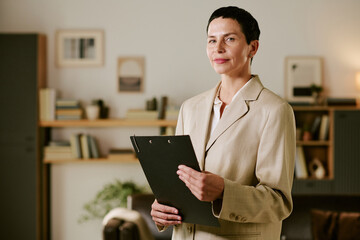 Portrait of middle aged Caucasian woman standing and holding clipboard, looking into camera with...