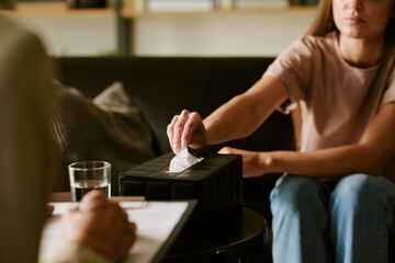 Young adult Caucasian woman reaching for tissue during therapy session with psychologist holding clipboard in foreground, illustrating psychological counseling process