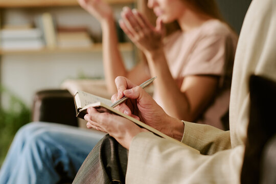 Middle aged Caucasian woman holding notebook and pen listening attentively while young adult Caucasian woman gesturing with hands during psychological counseling session
