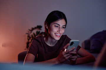 Young woman smiling while looking at her smartphone screen at home in low light, enjoying digital communication and apps