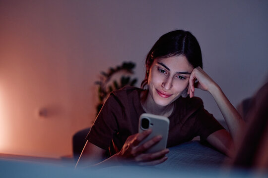 Young woman smiling, enjoying screen time on her smartphone while relaxing on a couch at home during the evening
