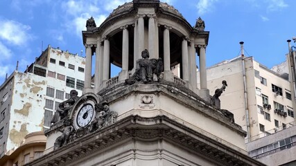 Close-up of ornate Neoclassical architecture in Rio de Janeiro's historic center. Features a detailed clock, dome, and classical statues set against a modern city background and blue sky.