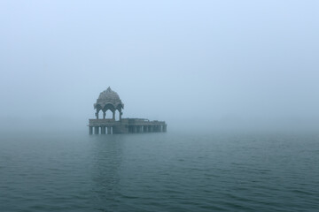 Obraz premium A serene early-morning view of a lone chhatri pavilion rising from the waters of Gadisar Lake in Jaisalmer, Rajasthan, surrounded by soft mist and tranquil reflection.