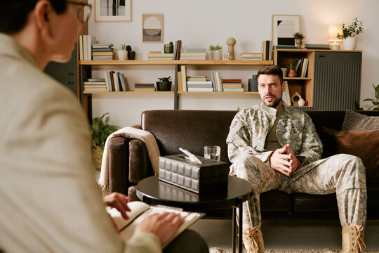 Caucasian young adult man in military uniform sitting on couch talking to middle aged Caucasian woman psychologist holding notebook during therapy session in office