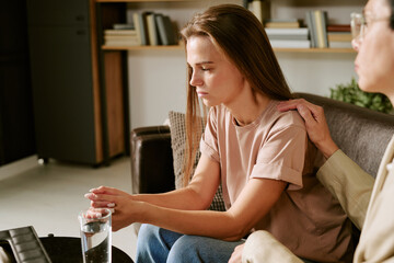 Caucasian teenage girl sitting on couch holding glass of water, looking down with serious expression, middle aged Caucasian woman gently placing hand on her shoulder during therapy session