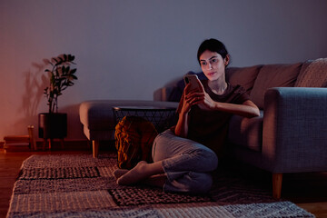Woman sitting on carpet in a dim lit living room holding smartphone, engaging with social media during evening downtime