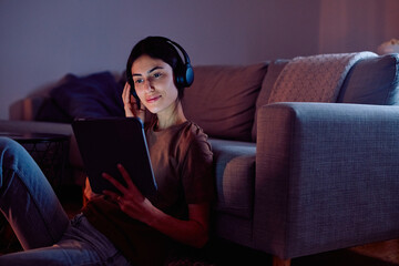 Woman wearing headphones and enjoying media on a tablet, relaxing on the floor at home in the evening