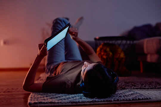 Young woman lying on floor with headphones, watching a screen on a digital tablet at night. Enjoying streaming content