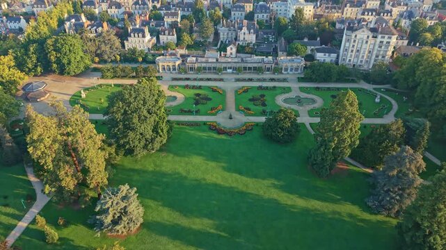 Drone pulls back over Thabor Park in Rennes, showing the bandstand, fountains, paths, French-style garden, surrounding buildings and sunrise light