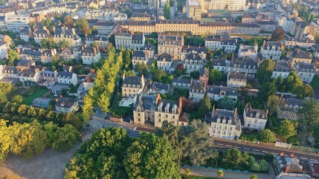 Drone shot moving backward above Thabor Park in Rennes, showing houses, trees, and the city under warm sunrise light.