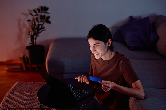 Young woman smiling on couch at night, holding credit card and using laptop for secure online shopping and digital payment convenience