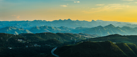 Aerial view of a winding highway running through vast green mountain range with electricity pylons under a golden sky at sunset