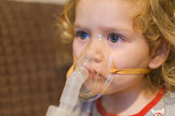 Little girl in medical mask doing inhalation at home. Child receiving medicine vapor from nebulizer. Bronchitis treatment.