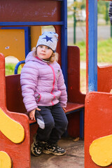 Young girl sitting thoughtfully on colorful playground bench in autumn. Child in purple jacket and star beanie on outdoor play structure during fall season.