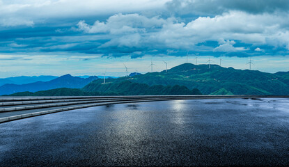 Empty asphalt road with beautiful mountain landscape and wind turbines generating clean electricity under a dramatic cloudy sky