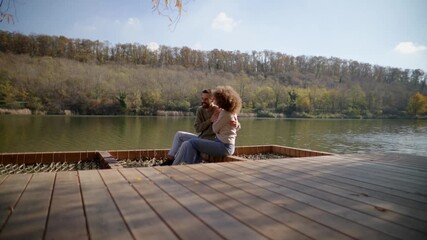 A couple enjoys a loving moment on a wooden dock, surrounded by autumn trees and calm water. Their smiles reflect joy and connection.

