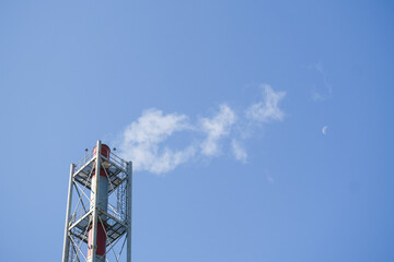 Industrial red smokestack emitting white vapor against clear blue sky with crescent moon. Factory chimney releasing steam on serene day for environmental themes