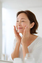 エイジングケア 中年女性が明るいバスルームで頬を触りながら素肌のうるおいを確認するスキンケアライフスタイル
