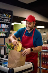 Happy supermarket cashier packing goods in paper bag at cash register.