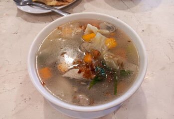 Indonesian Chinese dish, clear chicken soup with vegetables: carrots, cabbage, mushrooms, potatoes, tomatoes topped with fried shallots on a white bowl isolated on marble table.