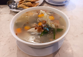 Indonesian Chinese dish, clear chicken soup with vegetables: carrot, cabbage, mushroom, potatoes, tomatoes topped with fried shallots on a white bowl isolated on marble table.