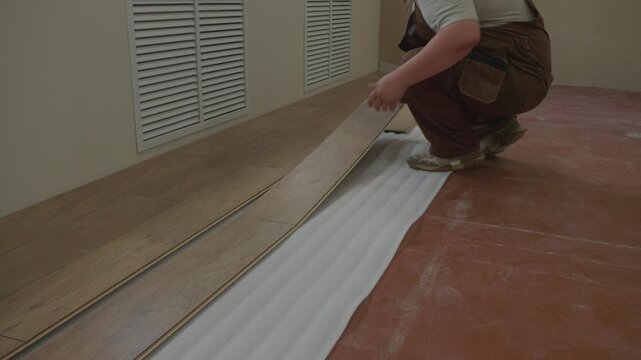 A hard-working construction worker in overalls carefully lays wooden laminate planks on a white underlay during a home renovation, meticulously adjusting every detail. Flooring installation.
