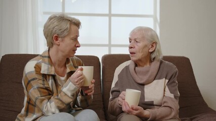 Two women elderly mother and middle aged daughter talking while sitting on couch in room over cup of tea. Attention and care for old people.