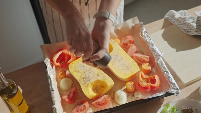 Person sprinkling butternut squash and vegetables at kitchen. Hands using pepper grinder to season fresh pumpkin and vegetables on baking tray. Concept of home cooking and healthy food preparation