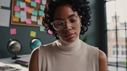 Woman sits at desk focused and thoughtful. Professional portrait shows eyewear and serious look. Workspace includes sticky note board. Brainstorm and planning vibe. Direct gaze for corporate profile.