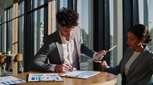 Review business document with man and woman. Colleagues discuss tablet and chart during office meeting. Teamwork and presentation planning in workspace. Partner gestures while peer notes. Growth now.