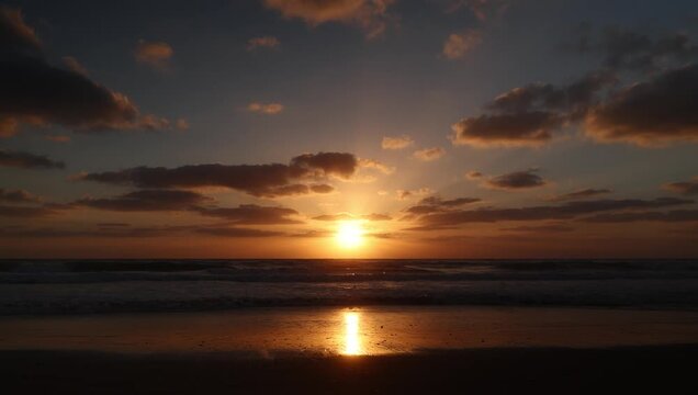 Golden sunset over the ocean with warm light reflecting on the wet sand, soft clouds framing the sun, and gentle waves creating a calm, atmospheric coastal scene.