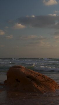 Dramatic  seascape with strong contrast, showing a large coastal rock in the foreground and dynamic waves under a cloudy sky.
