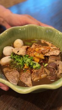 vertical video of Thai boat noodle bowl with rich broth meat slices herbs and steamed balls
