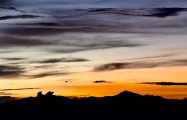 Dramatic sunset with intense orange and yellow hues piercing through layered clouds, silhouetting rugged mountain peaks. Soft gradient sky transitions to deep blue. 