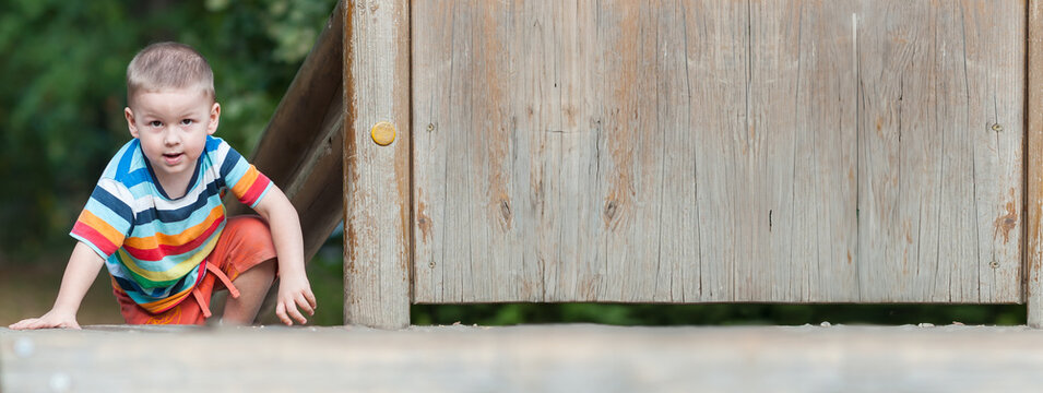 A cheerful young boy in a colorful striped shirt crawls near a wooden playground structure. He explores his surroundings with curiosity while enjoying the warm weather outdoors. Place for text
