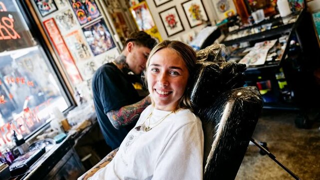 Smiling woman in tattoo studio relaxing in chair while artist prepares tools, candid lifestyle scene of courage, self‑expression and modern body art culture &mdash; cinematic stock video in authentic style,